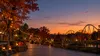 AI-generated editorial image of an autumn amusement park at twilight with a distant roller coaster, seasonal lights and pumpkins.