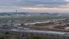 An airport runway complex operates beside a large graded construction corridor under gray morning skies.