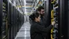 Two data-center engineers inspect unbranded optical network connections among rows of server cabinets.