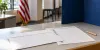 Unmarked ballot envelope, blank ballot and pencil on a civic office counter beside a privacy screen