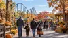 Families walk through an autumn amusement-park midway decorated with pumpkins, with a roller coaster in the distance.