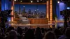 A late-night television studio with an empty host desk, guest chair, cameras and audience before a city skyline