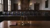 An empty courtroom with 12 jury chairs, one separated from the others, and balanced scales of justice in the foreground.