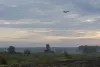A distant fighter aircraft above a radar installation in a Baltic countryside landscape