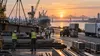 An unbranded Louisiana construction and logistics worksite at dawn with cranes, freight equipment and workers shown only as distant silhouettes in a restrained editorial scene.