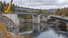 Editorial illustration of steel bridge girders being placed over a Maine river beside a temporary bridge in autumn
