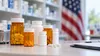Unbranded prescription bottles and tablets on a pharmacy counter with a blurred American flag in the background.