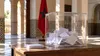 Transparent ballot box with unmarked ballots and a Moroccan flag in a civic interior