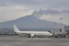 An unbranded passenger aircraft sits beneath a hazy sky with Mount Etna visible beyond the airport.