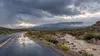 Storm clouds over a rain-soaked New Mexico road beside a flowing desert arroyo.
