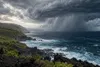 Editorial image of dense storm clouds and heavy rain approaching a volcanic Hawaiian coastline.