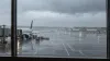 Rain streaks terminal windows overlooking aircraft on a wet airport apron during a coastal storm.