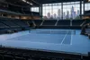 Conceptual view of an empty blue tennis court inside a modern arena with a generic city skyline beyond the windows.
