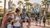 A multigenerational family checks a phone while a child carries a blank sketch pad outside a generic palm-lined Central Florida theme park.