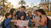 A multigenerational family reviews a blank itinerary and phone at a generic palm-lined Central Florida theme park decorated for fall.