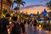 Visitors check a smartphone while walking through a tropical Central Florida theme-park promenade near sunset.