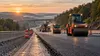Fresh asphalt, road rollers and safety cones on a Pennsylvania highway work zone against wooded hills at dawn.
