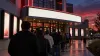 Moviegoers line up outside a contemporary cinema beneath blank illuminated marquees at dusk.