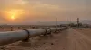 A large oil pipeline crosses a Saudi desert landscape toward an industrial pumping station at sunset.