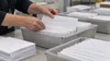 Adult hands sort plain sealed ballot-style envelopes into neutral postal trays at an election-administration worktable.