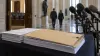 A sealed briefing folder and stacks of documents sit beside microphones in a congressional corridor.