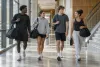 Four college student-athletes carrying unbranded gear bags through a university athletics facility