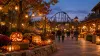 An illustrative autumn theme park walkway with glowing pumpkins and a roller coaster at dusk.