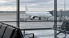 AI-generated illustrative view of an unbranded passenger jet beyond an airport terminal window; not a photograph of a specific aircraft or event.