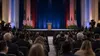 A generic civic convention hall prepared for a major political address, with an empty lectern and press cameras