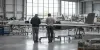 Two technicians inspect unbranded unmanned-aircraft components on a neutral aerospace factory floor.