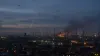 Distant drones and searchlights above an oil refinery at predawn, with smoke rising from an illuminated industrial unit