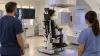 A patient seated upright in a radiotherapy treatment room while two clinicians monitor positioning.