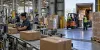 Workers process packages on a manufacturing line beside an active warehouse loading dock
