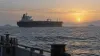 An unmarked oil tanker crosses calm Persian Gulf waters at dawn with distant naval vessels on the horizon.