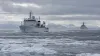 An unmarked white research ship crosses broken Arctic sea ice while a distant cutter monitors from offshore.