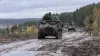 Armored vehicles travel along a muddy training road in a Baltic forest as soldiers coordinate nearby.