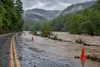 Floodwater covers part of a rural Appalachian road near wooded hills, with traffic cones marking the hazard under storm clouds.