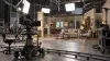 An unbranded television camera and production equipment facing a constructed interior set on a professional soundstage
