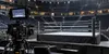 An empty professional wrestling ring in an indoor arena with a broadcast camera positioned at ringside.