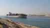 Un buque portacontenedores sin distintivos atraviesa el Canal de Suez a la luz del día, pasando junto a la infraestructura portuaria situada al borde del desierto