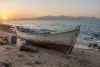 An unmarked wooden passenger boat beside water containers and blankets on an arid shoreline at dawn.