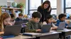 Elementary students use classroom laptops while a teacher observes in a generic public-school setting.