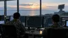 Military radar operators monitor unlabelled tracking displays in a coastal operations room overlooking the sea at dawn.