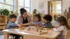 Preschool teacher guides four children playing with wooden learning materials in a bright early-learning classroom.