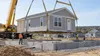 Operários guiam uma secção pré-fabricada de uma casa em direção a uma fundação de betão num estaleiro de obras residencial.