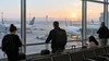 Travelers watch parked commercial aircraft from an airport terminal at dawn.