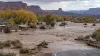 Illustrative view of muddy floodwater surrounding homes and a road in a high-desert community as rescue boats cross the water.