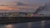 A symbolic editorial image of an airport terminal at dusk with emergency lights and smoke rising nearby.