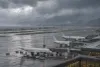 Um tranquilo pátio de aeroporto na costa do Pacífico sob nuvens de tempestade carregadas, com aeronaves de passageiros comuns estacionadas perto de um terminal moderno e a chuva a cair com intensidade na pista.