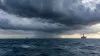 Imagem editorial gerada por IA de nuvens escuras de tempestade tropical sobre as águas do Golfo, com duas plataformas de energia offshore distantes.